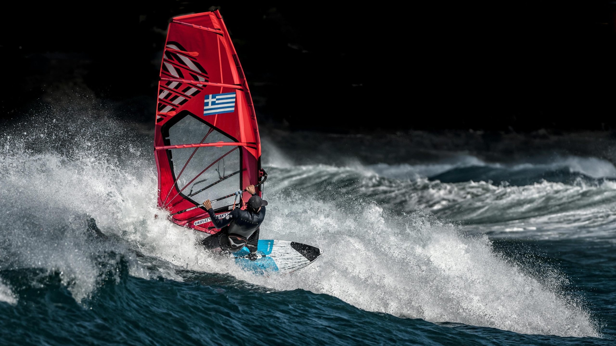 A thrilling capture of a windsurfer riding a wave with a Greek flag sail, showcasing extreme sports action.