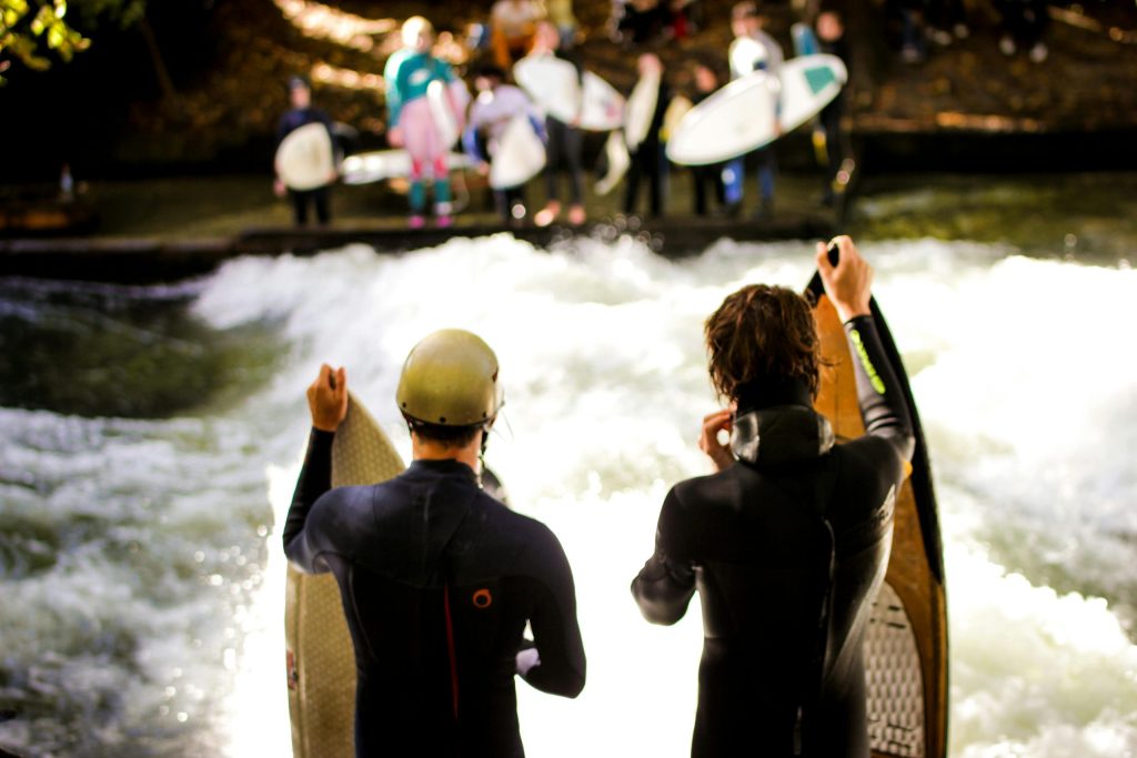 Surfers prepare to ride on river waves, surrounded by an energetic group.